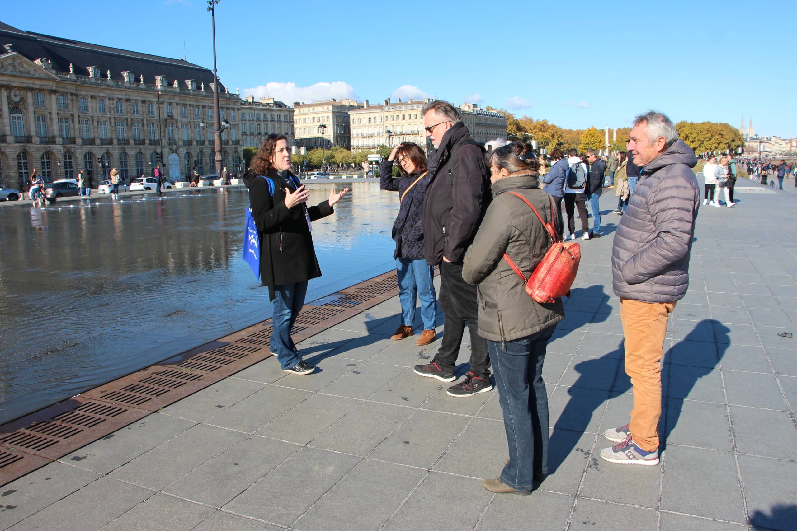 Guide expliquant un lieu à quatre touristes devant le Miroir d'eau de Bordeaux, par temps clair.