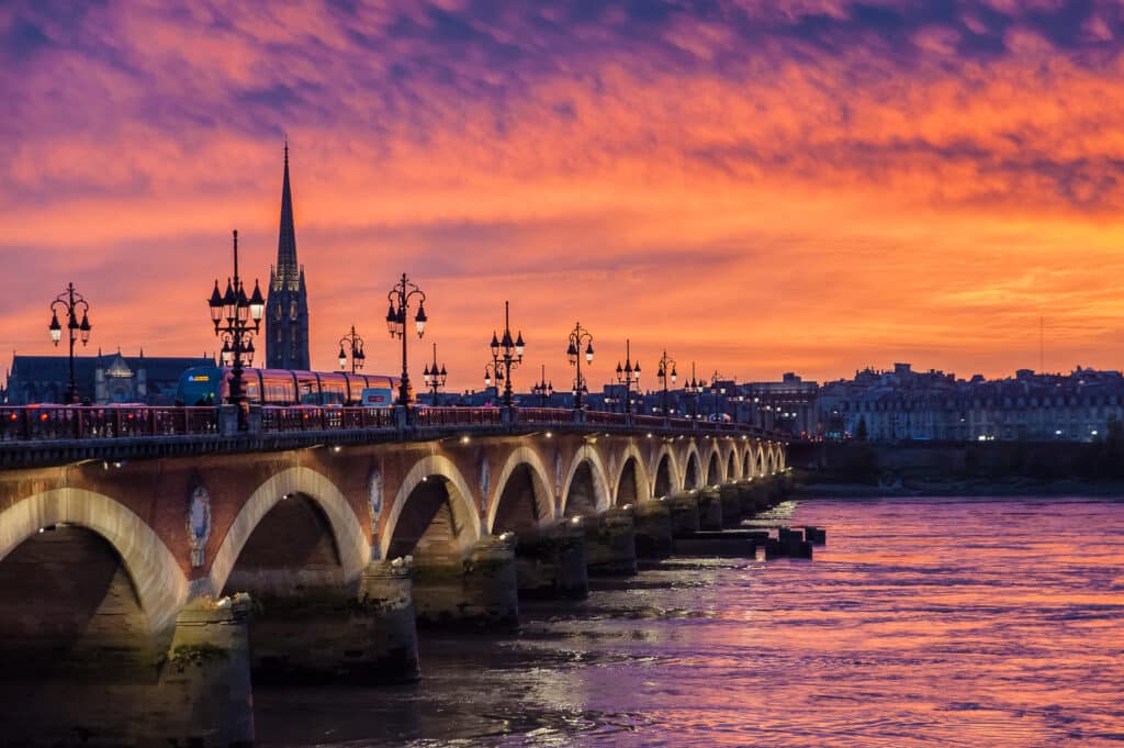 Coucher de Soleil Bordeaux : Pont de Pierre et Tramway Pont de pierre illuminé au coucher du soleil flamboyant sur la Garonne, tramway rouge et flèche de cathédrale en arrière-plan.