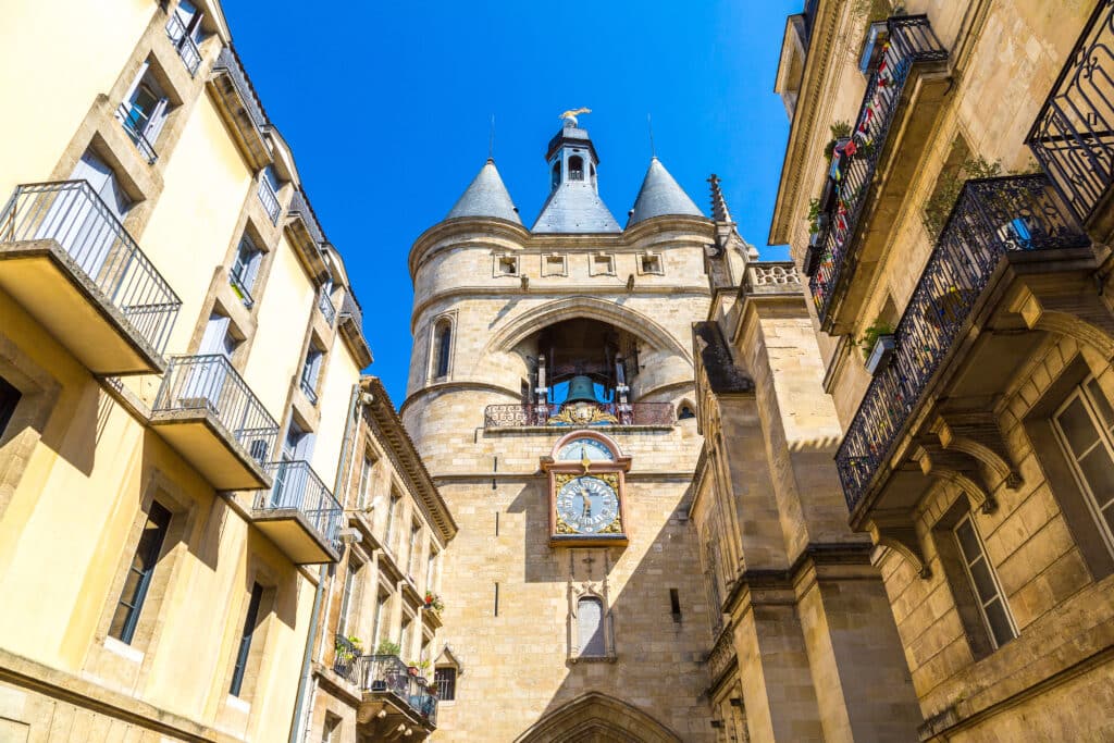 La Grosse Cloche de Bordeaux : façade et horloge Grosse horloge de la Grosse Cloche de Bordeaux, encadrée par des façades ocres à balcons en fer forgé, sous un ciel bleu vif.